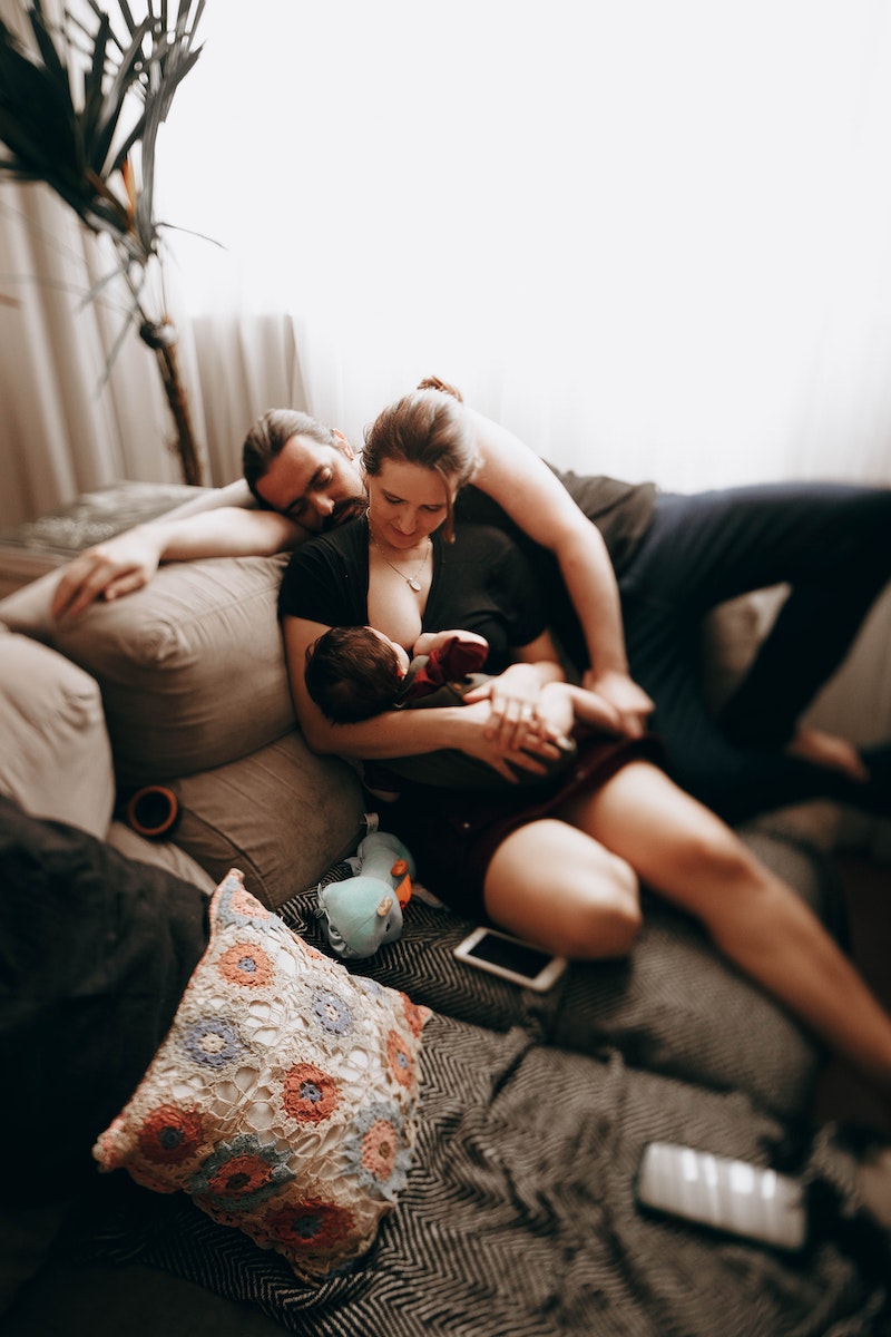 Caring mother breastfeeding little baby while sitting on comfortable sofa near father while spending time together at home in living room