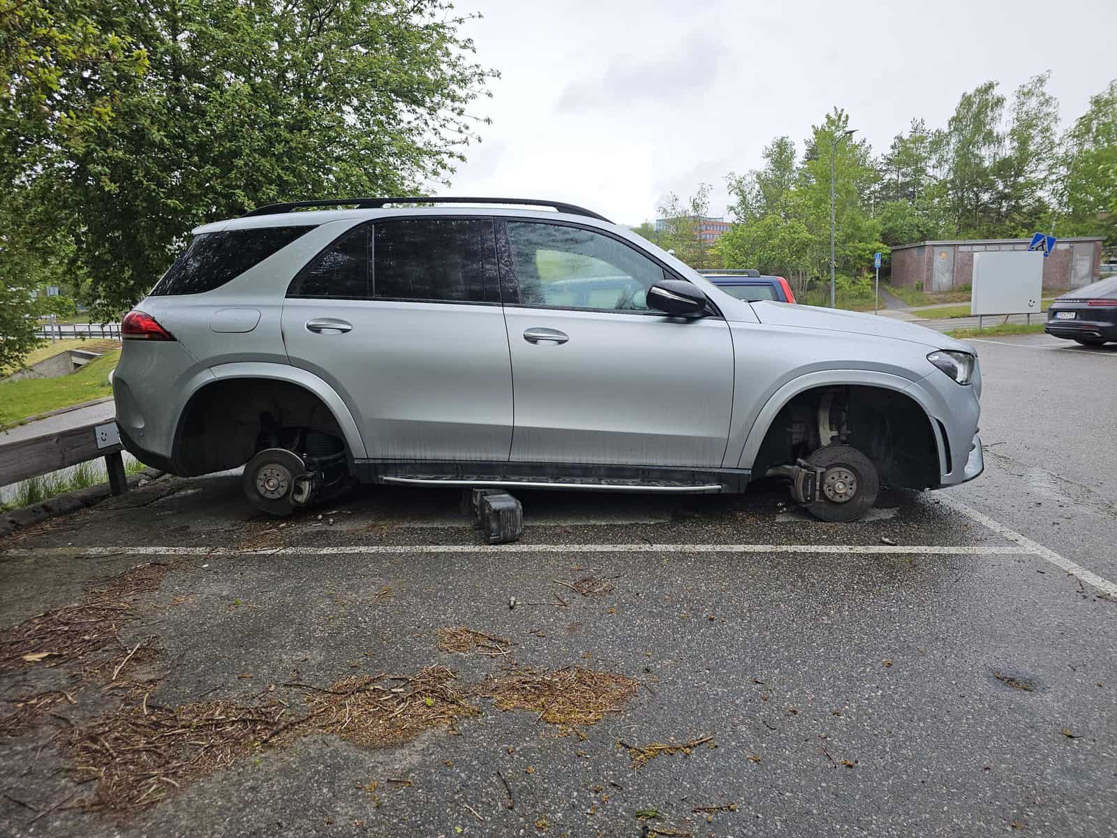Silver SUV parked in a residential lot with all four wheels stolen, sitting on small wooden blocks with exposed brake rotors—appears to be the victim of recent vandalism or theft.