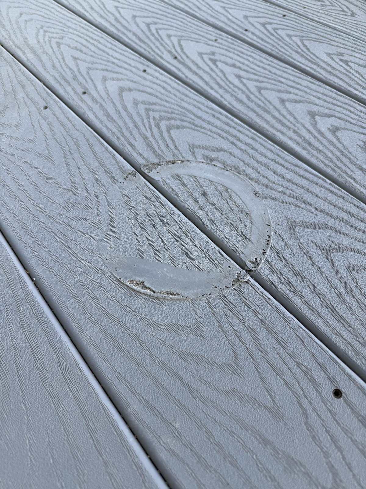 Melted plastic ring fused to gray composite decking, likely caused by placing a hot pot or pan directly onto the surface—burn mark is circular with distorted plastic edges.