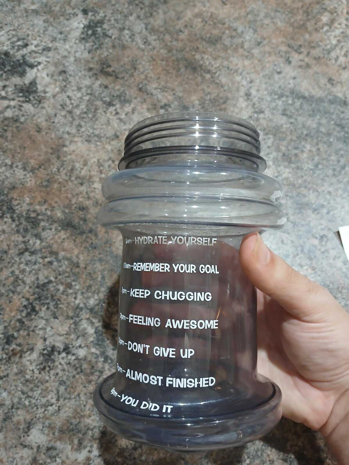 Motivational water bottle with time-based encouragements from 9 a.m. to 6 p.m., held in a person’s hand on a granite countertop—bottle is crumpled and deformed, indicating it was likely damaged or melted.