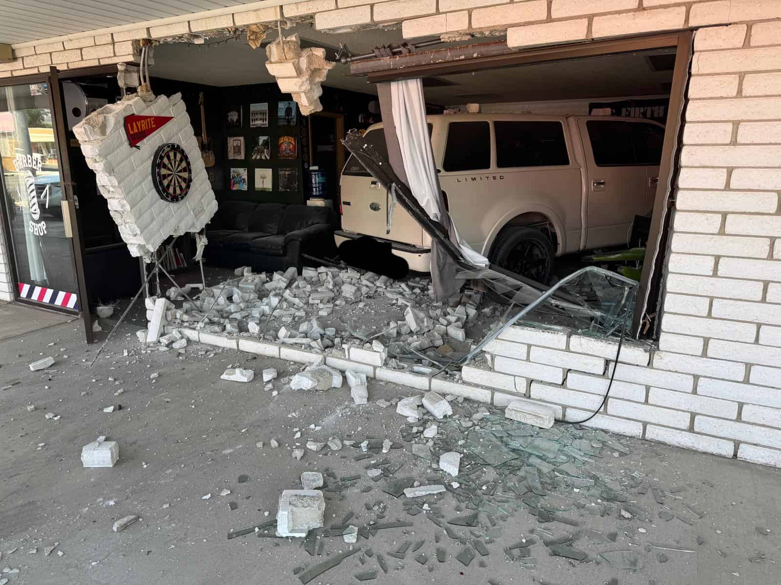 White SUV crashed through the front wall of a barbershop, collapsing a large section of brick and glass into the interior—dartboard, posters, and couch inside remain intact amid rubble.