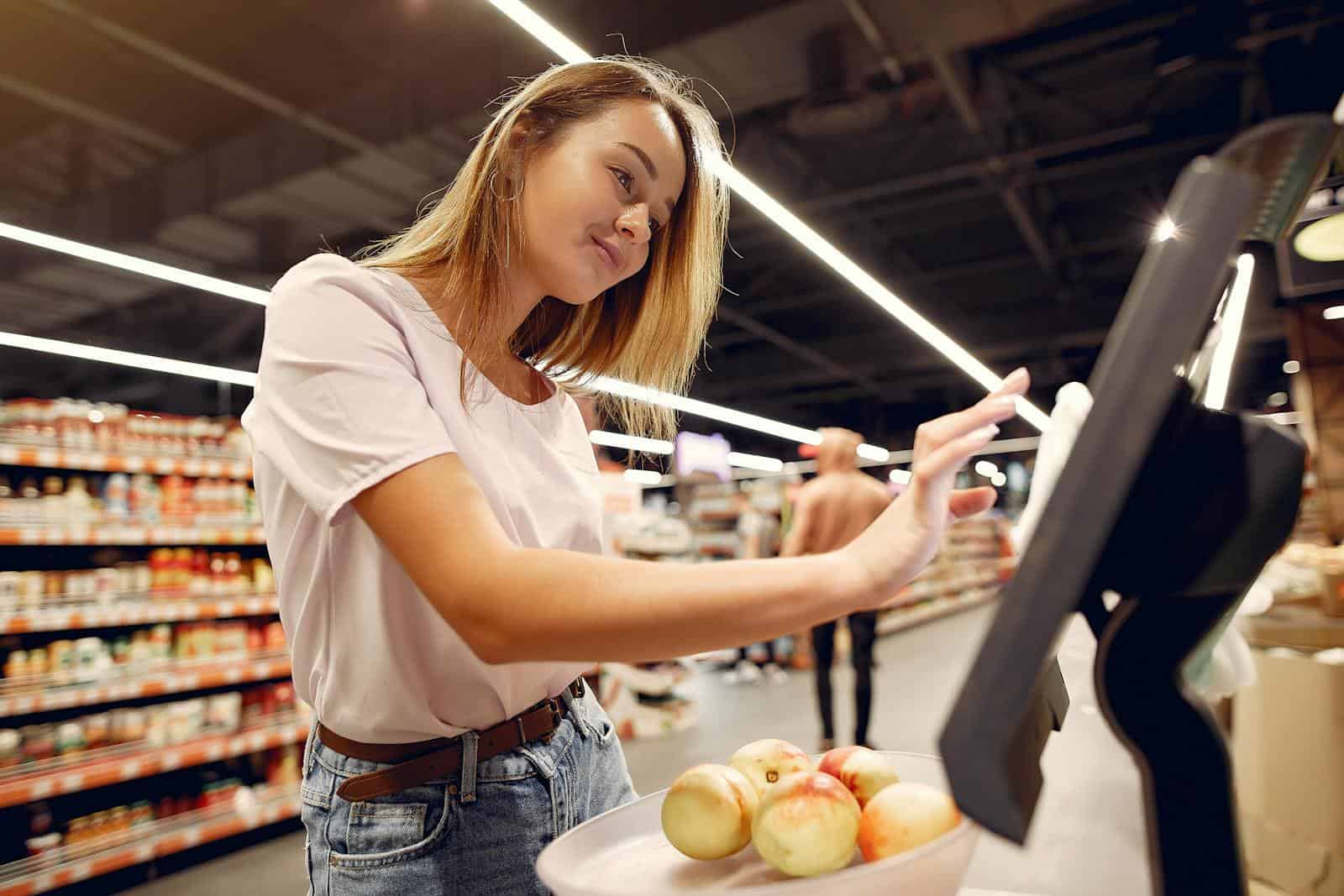 Side view of young woman in trendy clothes weighing peaches on scales while shopping in supermarket during purchase food