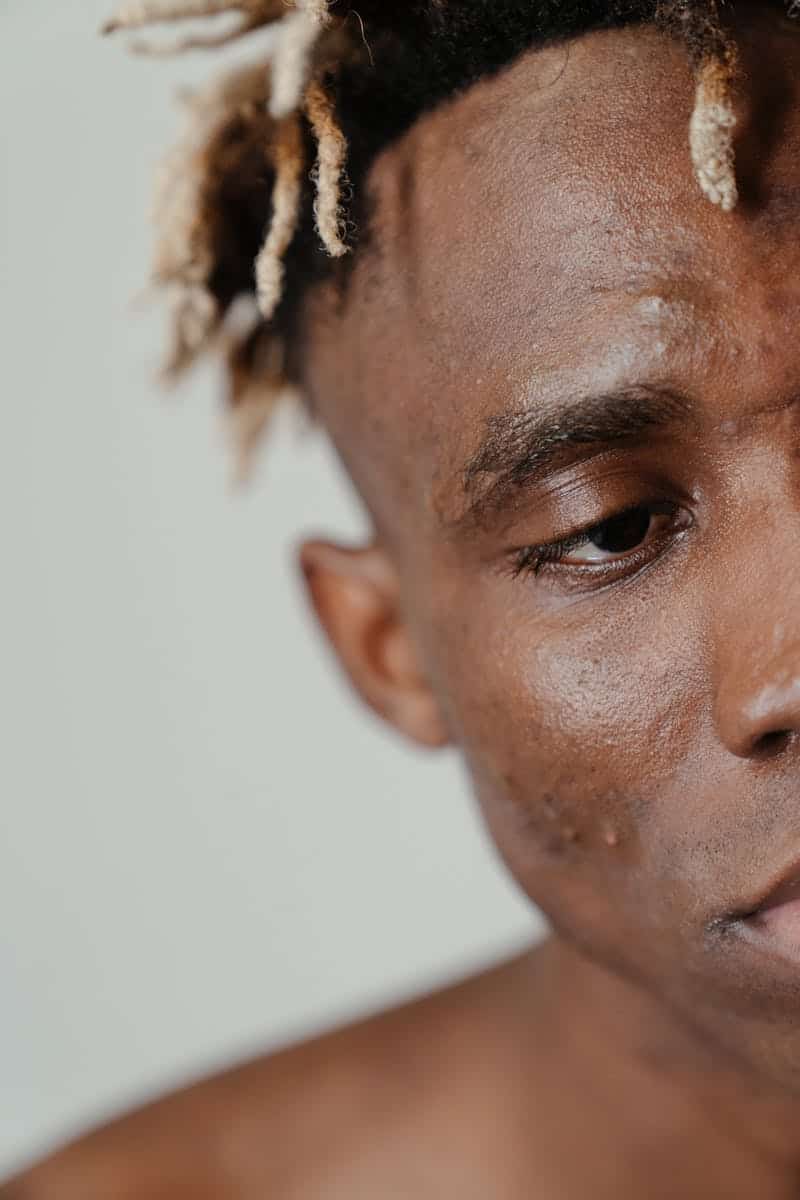 Close-up portrait highlighting the skin texture and dreadlocks of a shirtless young man.