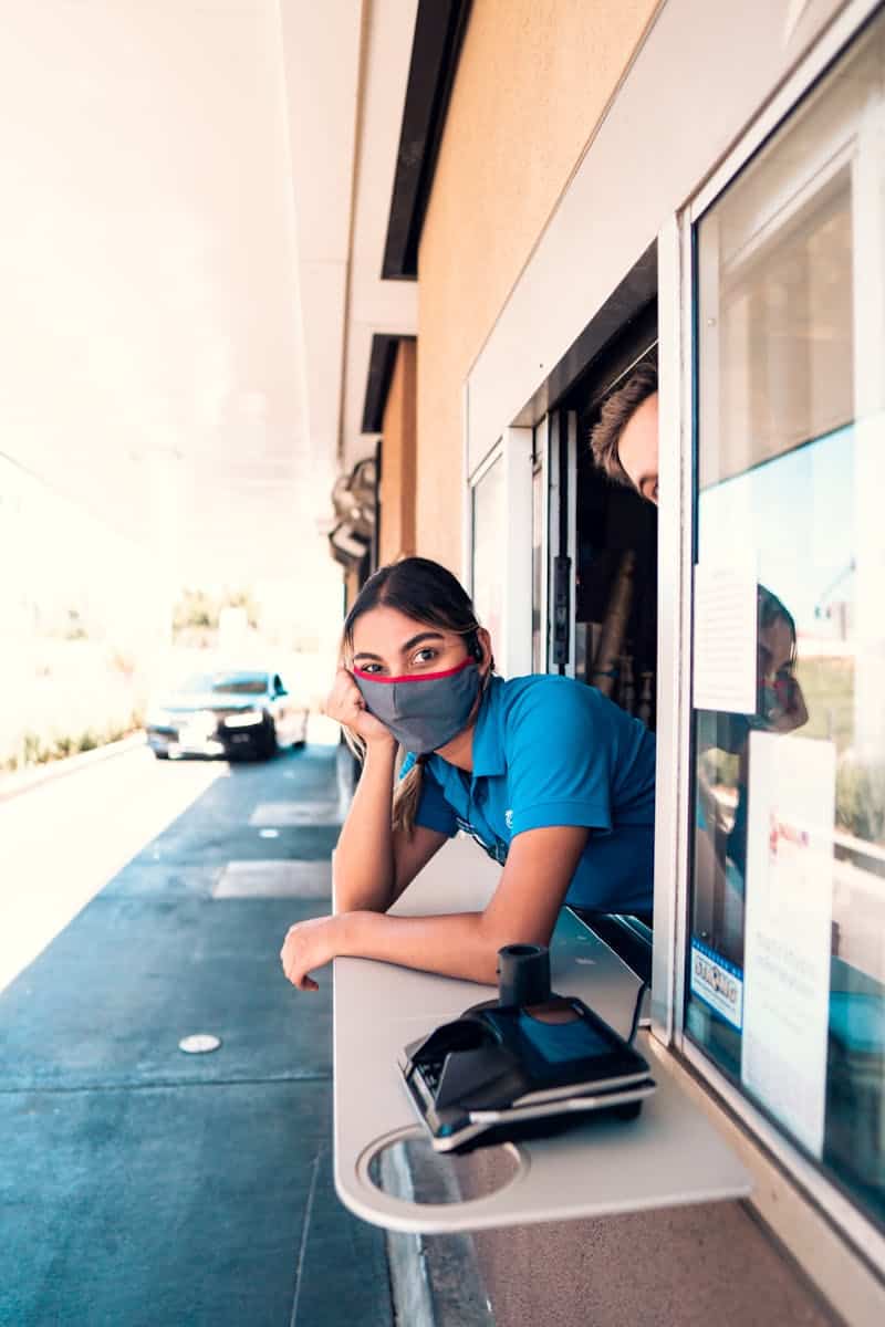 Woman in mask at fast food drive-thru window, serving customer with contactless payment.