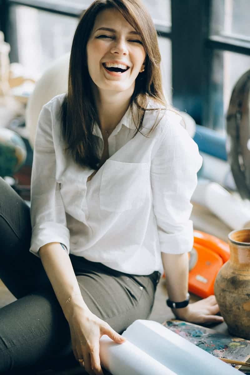 A woman in casual attire laughs joyfully while sitting indoors, exuding happiness and carefree energy.