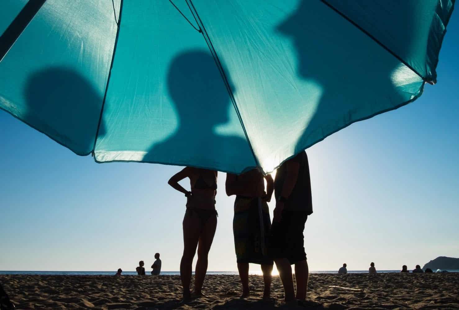 Group of people under a teal beach umbrella on the sandy shoreline.