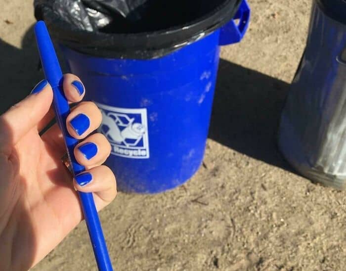 Blue pen held by a hand with matching blue painted nails, near a blue recycling bin outdoors.
