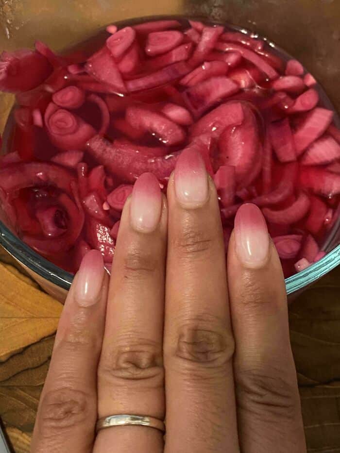 Brightly colored pickled onions in a glass bowl, close-up of a hand with a wedding ring.