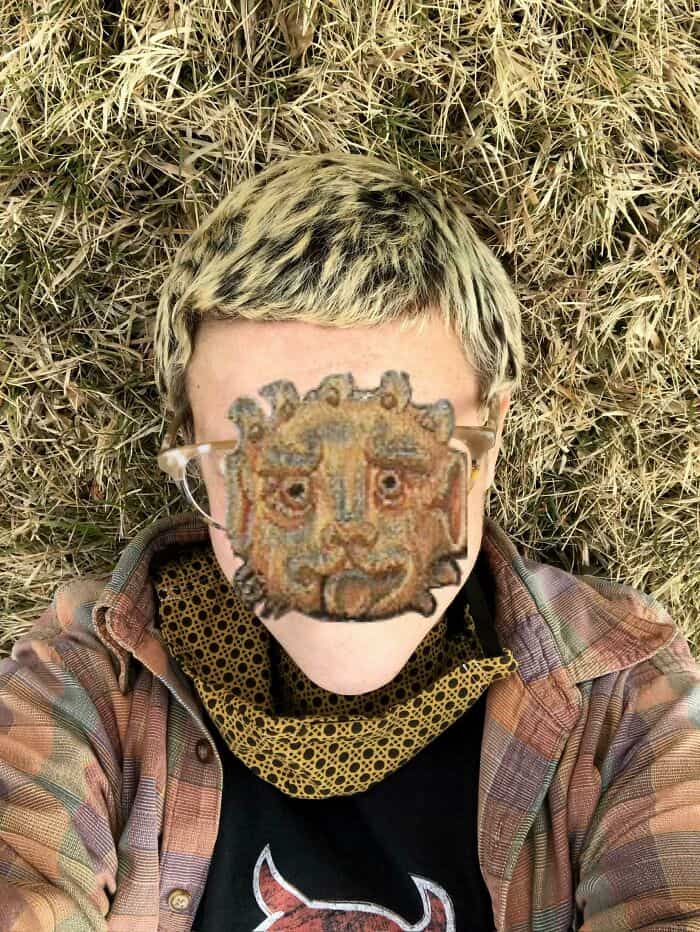 Person wearing a lion face mask lying on dry grass, casual clothing, and glasses.