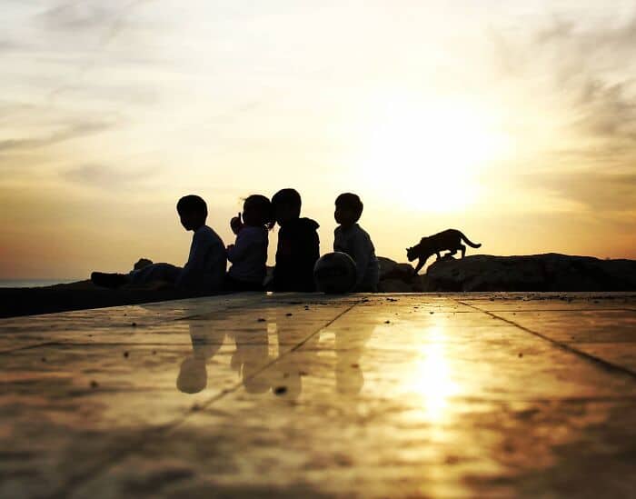 Children sitting on the ground at sunset with a dog running nearby.
