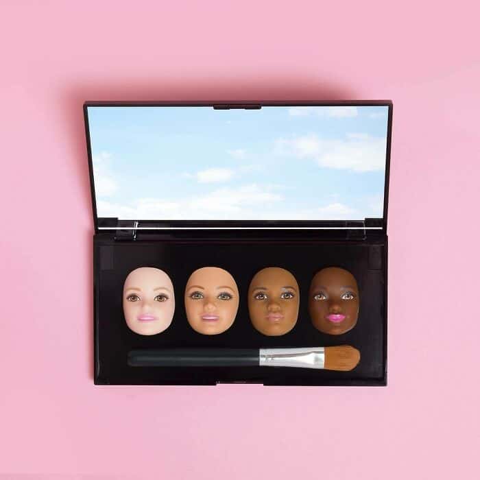 Makeup brush and four diverse doll heads in a black case on pink background.