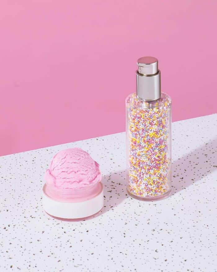 Pink glitter spray bottle with colorful confetti-like sprinkles, and a pink cream container, on a sp.
