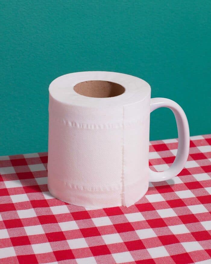 Toilet paper roll mug on checkered tablecloth with teal background, blending humor and surrealism.