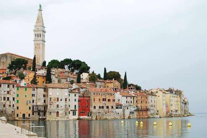 Vibrant seaside city with colorful buildings, a tall historic tower, and calm water reflecting the s.