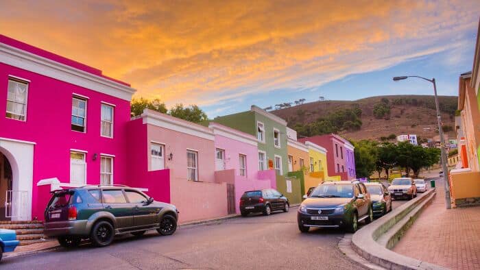 Vibrant, colorful houses on a city street during sunset, with parked cars and a scenic hillside in t.
