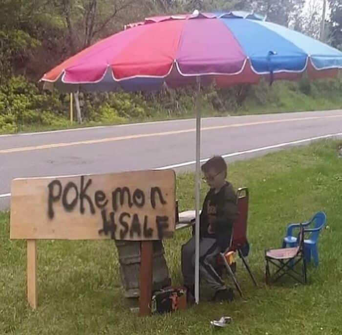 Child sitting at Pokémon sale stand under colorful umbrella by roadside.
