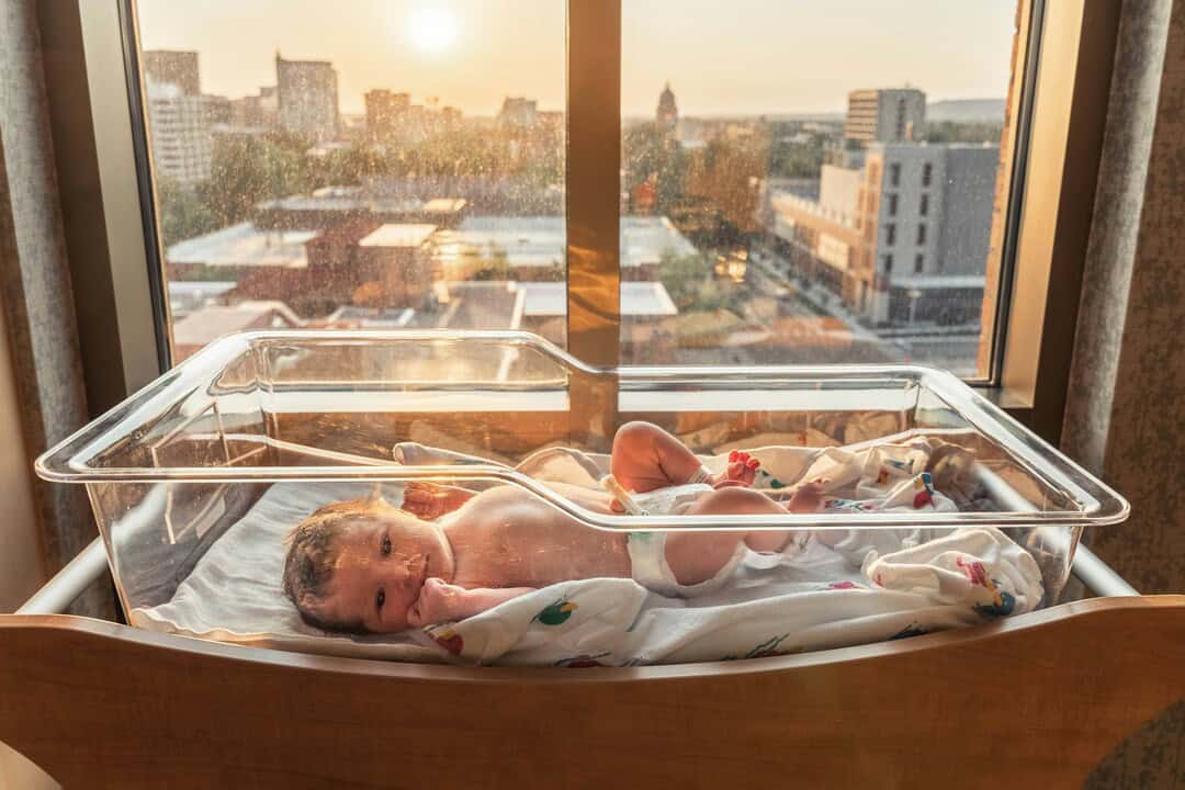 Baby in hospital bassinet near window with city view at sunset.