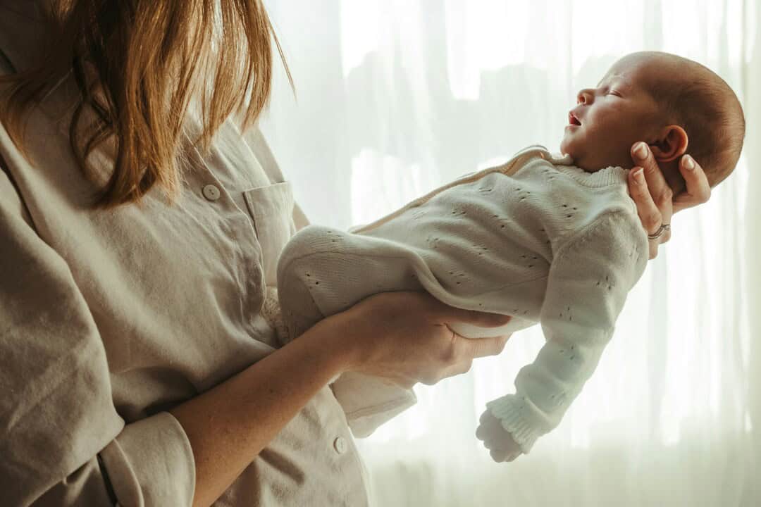 Baby being held by a woman in front of a bright window.