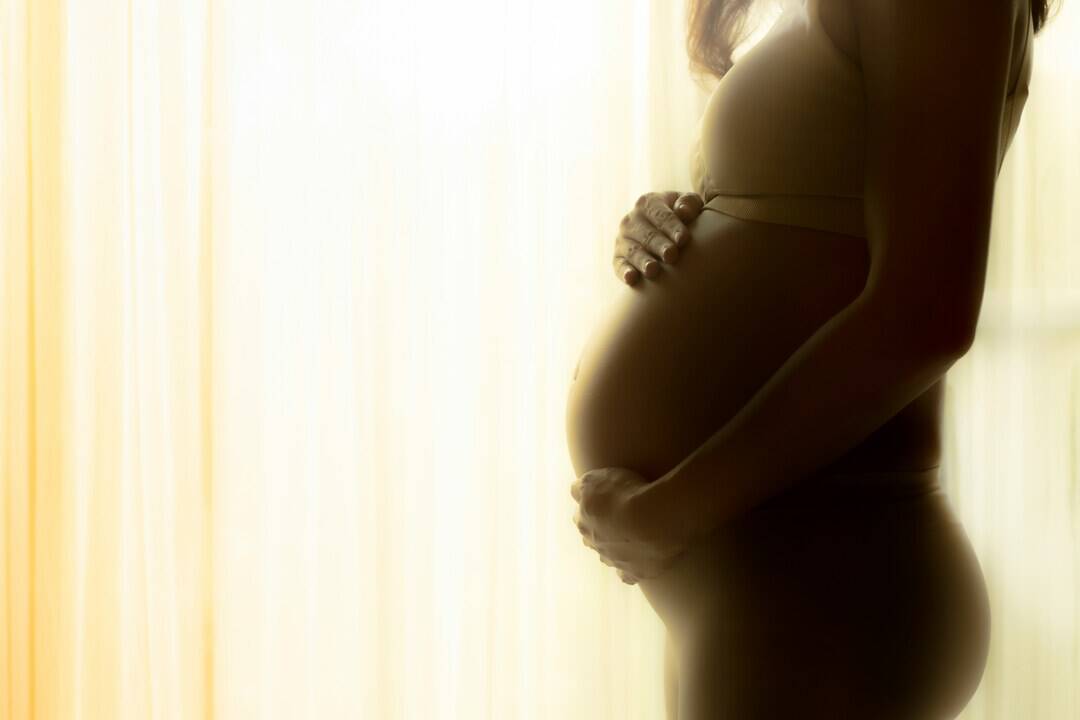 Pregnant woman holding her belly, backlit by soft natural light, highlighting pregnancy and maternal.