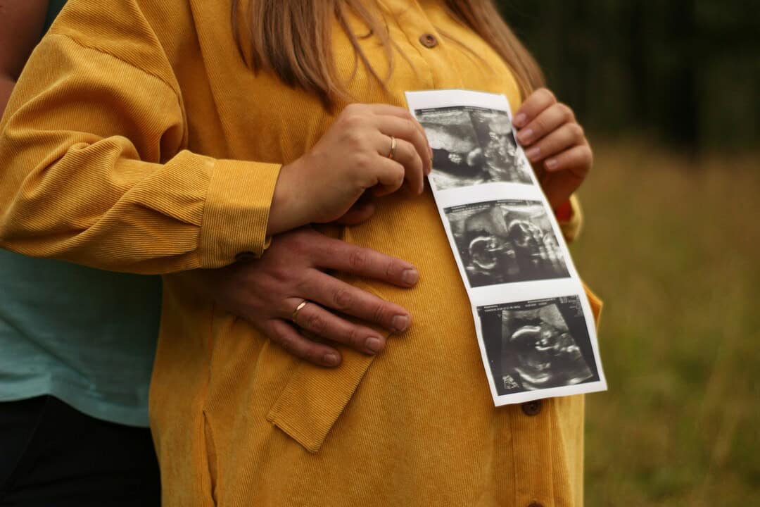 Pregnant woman holding ultrasound images with partner outdoors.