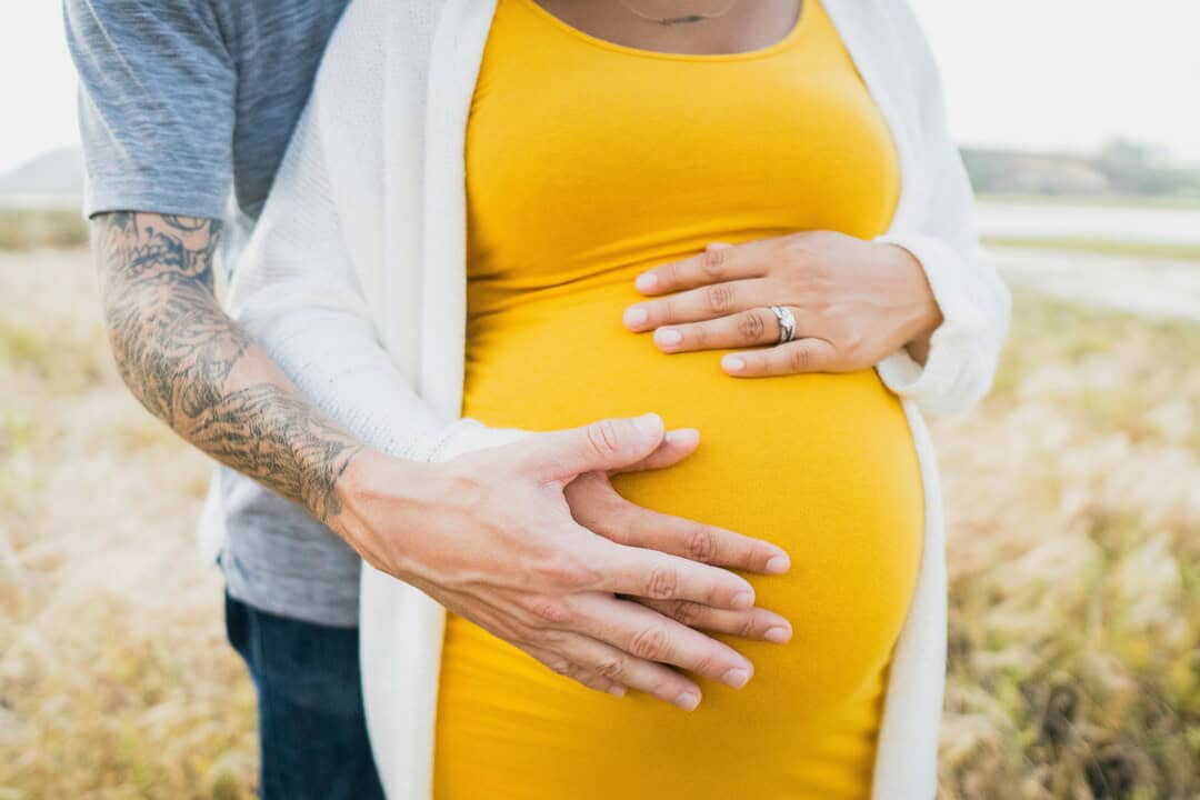 Close-up of pregnant woman with partner's hands on her belly in outdoor setting.