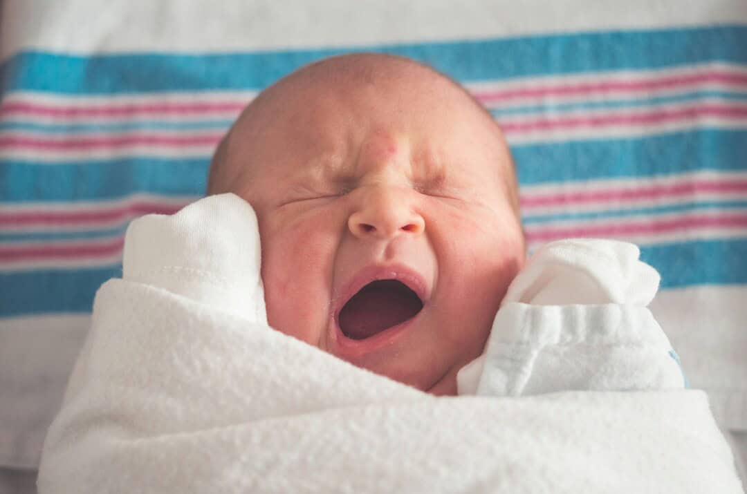 Baby yawning with eyes closed, wrapped in a white hospital blanket, on a colorful striped background.