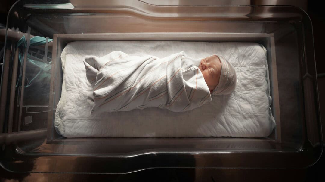 Baby swaddled in blanket inside neonatal incubator.