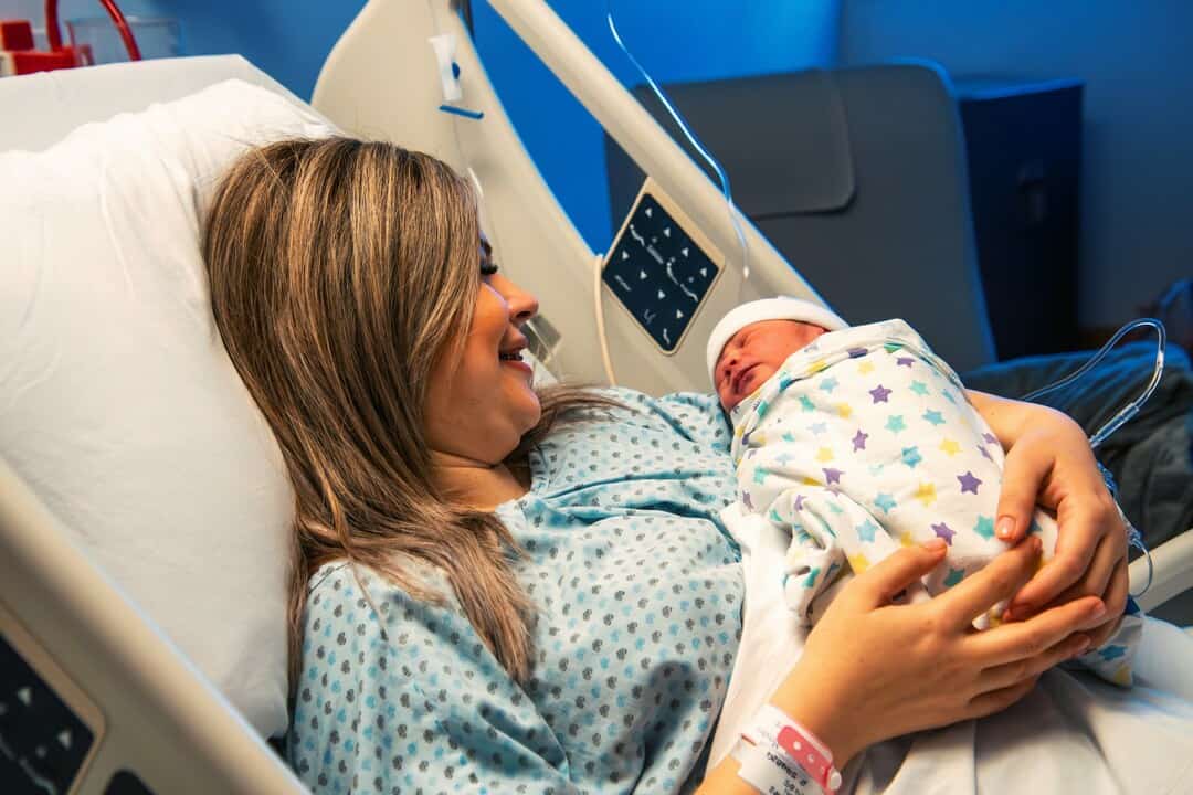 Mother holding newborn baby in hospital bed, smiling and bonding after childbirth.