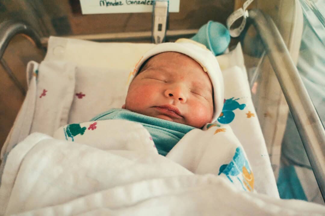 Newborn baby sleeping peacefully in hospital crib with medical equipment nearby.