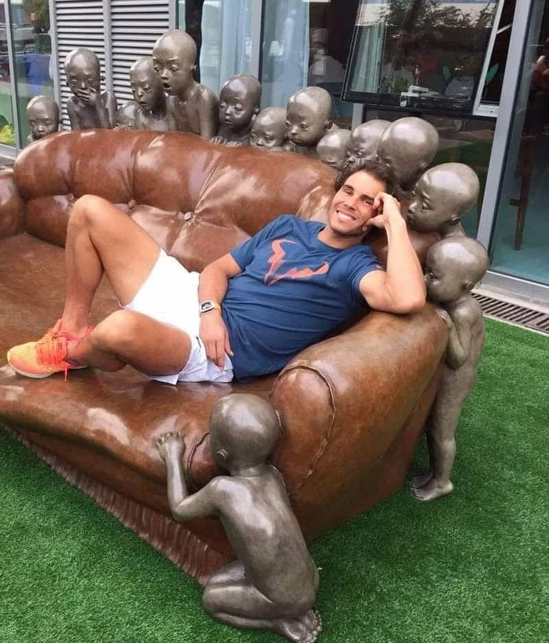 Relaxed man lounging on leather sofa with sculptures of children, outdoor setting.