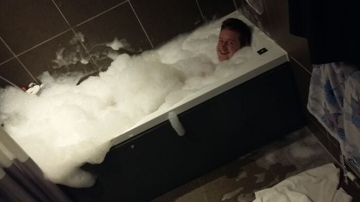 Man smiling and relaxing in a bubble-filled bathtub at home.