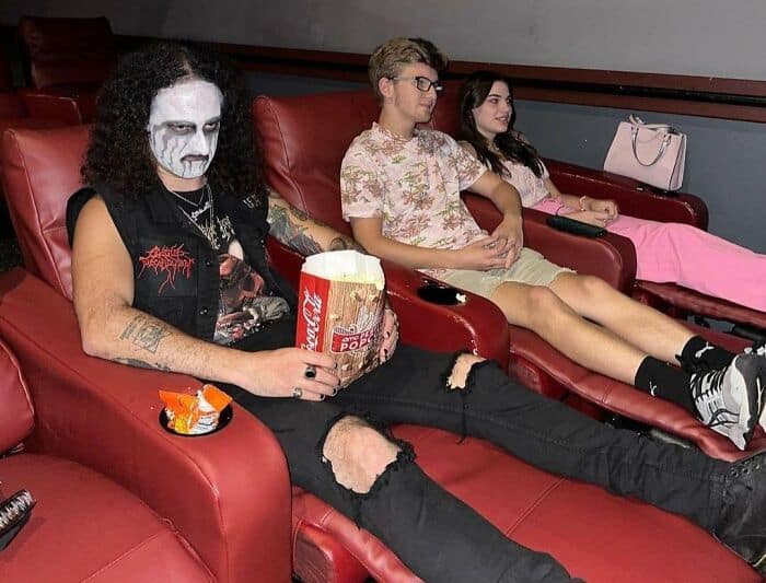 Men sitting in theater seats, one with face paint, casual clothing, and snacks.
