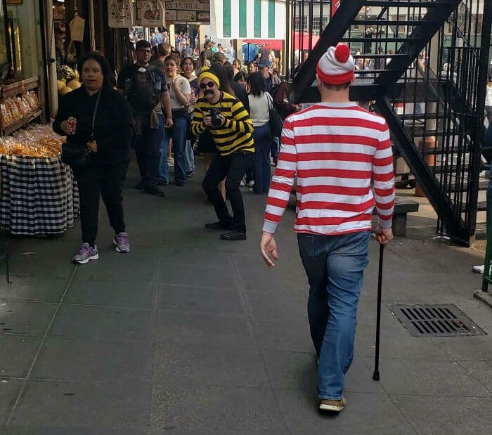 Man dressed as Waldo with cane walking through crowded outdoor market.