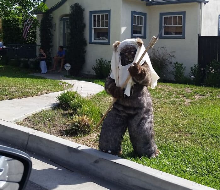 Man dressed as Bigfoot in costume holding a stick outside house.