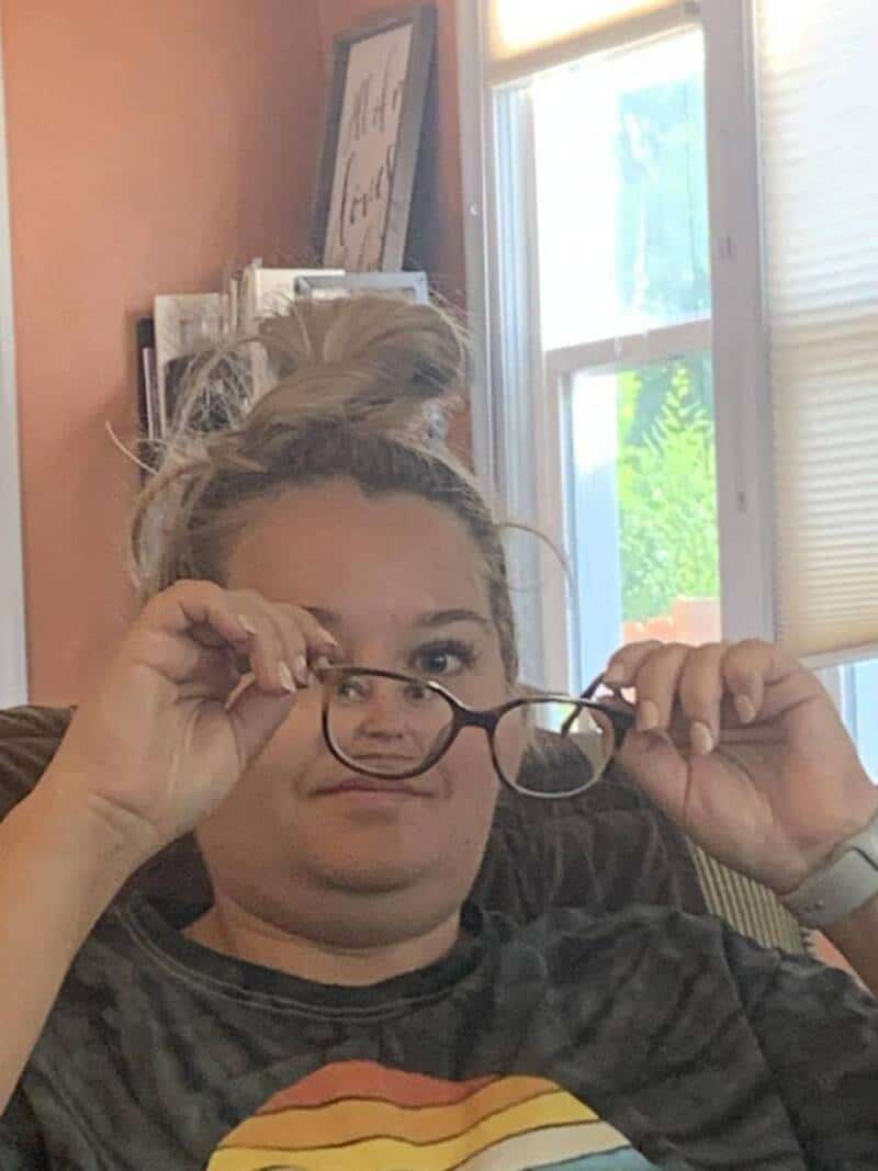 Woman holding glasses, making a humorous face indoors with natural light from a window.