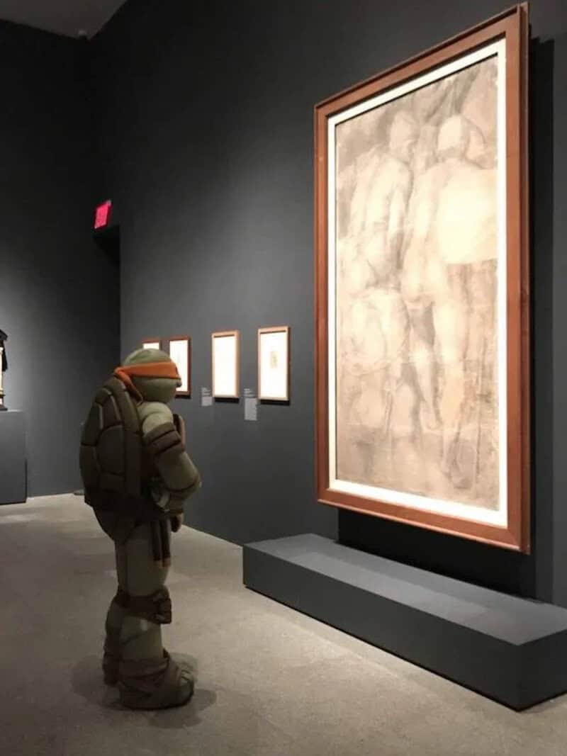 Astronaut in space suit looking at framed artwork in a gallery setting.