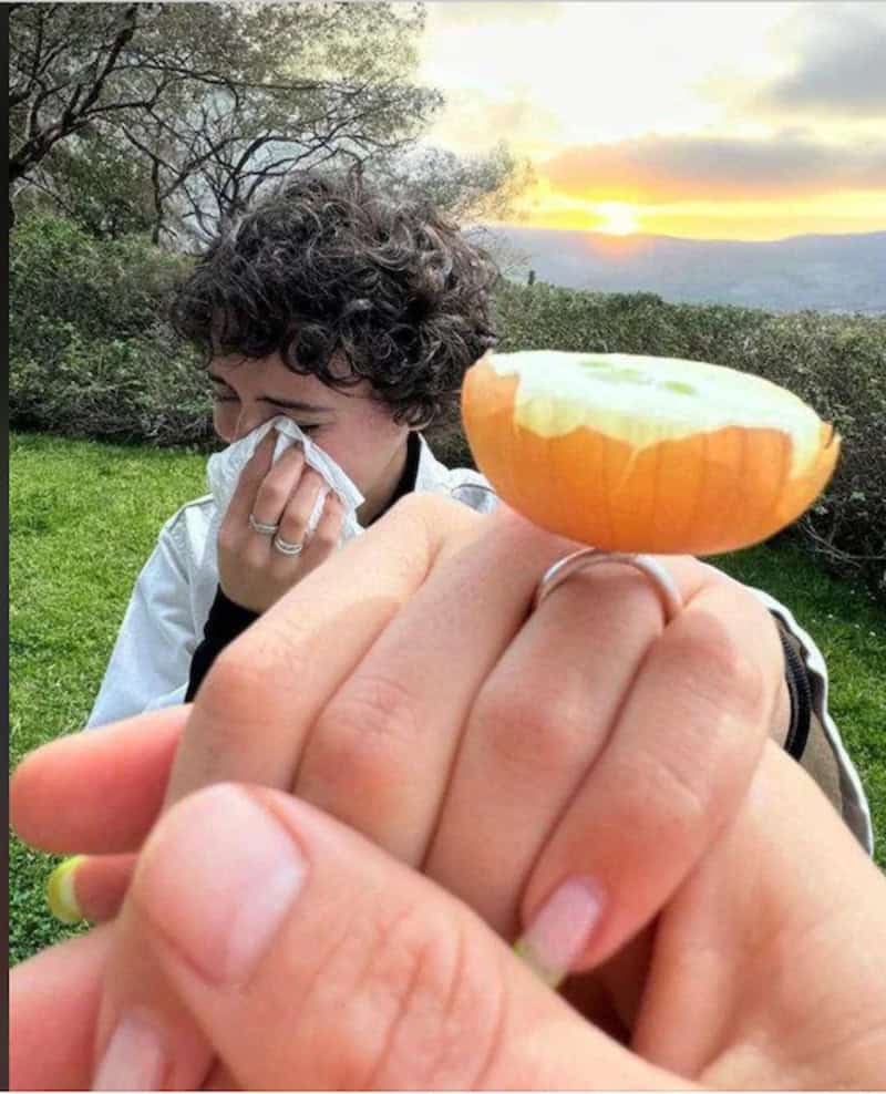 Close-up of a hand holding a cracked egg with a person in the background wiping their face, set outd.