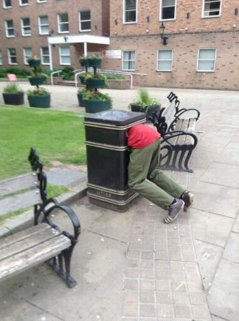 Person with head in trash can on city sidewalk, humorous scene.