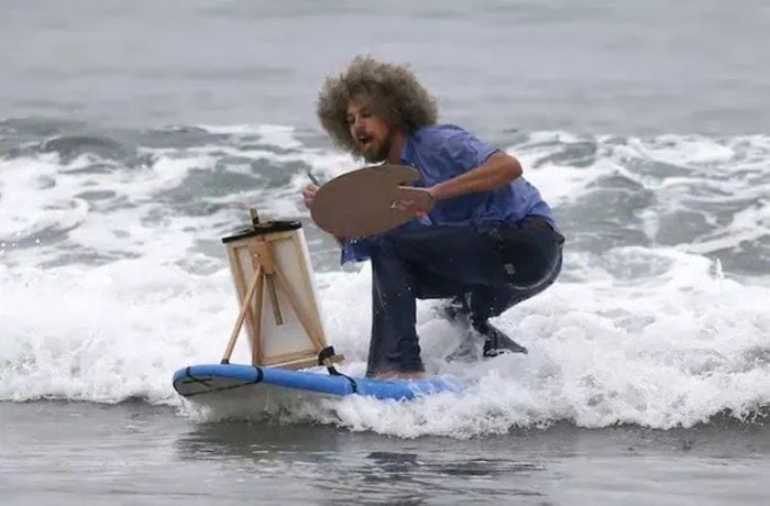 Man with curly hair painting on a surfboard at the beach.
