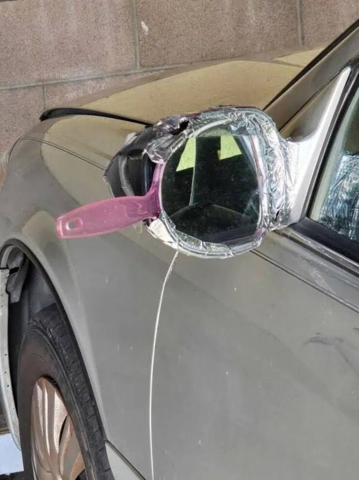 Car side mirror covered with aluminum foil and pink handle, taped to the vehicle.