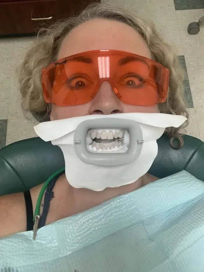 Woman with dental mouth prop, orange safety glasses, and dental bib in dental chair.