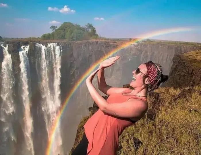 A woman with sunglasses and a headscarf appears to be holding a rainbow with her hands near a waterf.