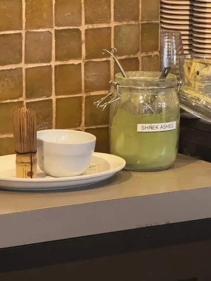 SHREK ASHES in a glass jar with a spoon, on a counter with a white bowl and a brush, against a tiled.