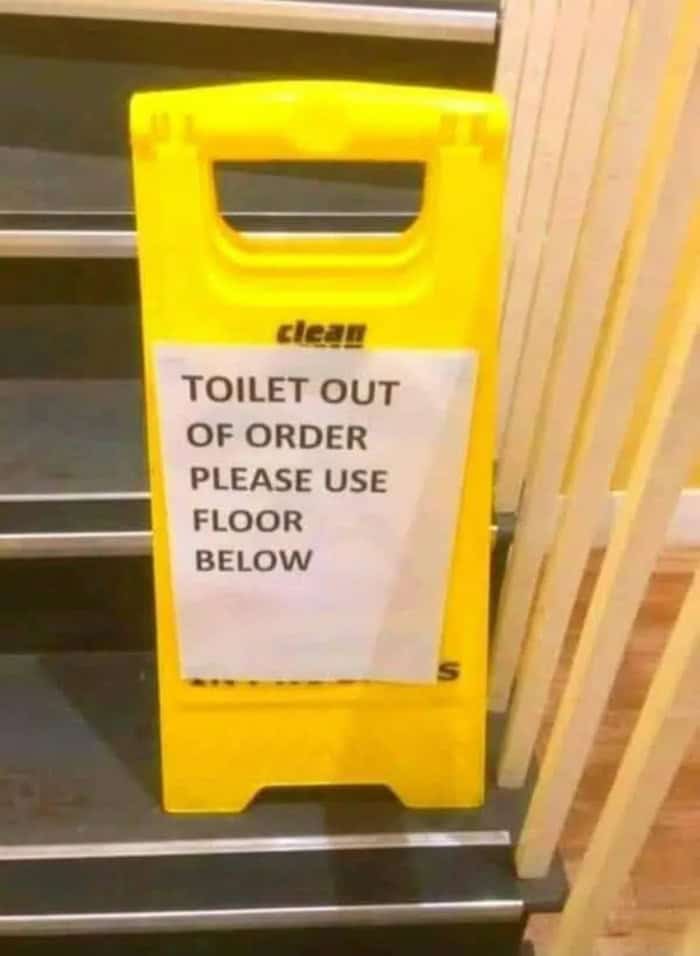 Out of Order Toilet Sign, Please Use Floor Below, Yellow Stand, Indoor Setting, Near Wooden Railing.