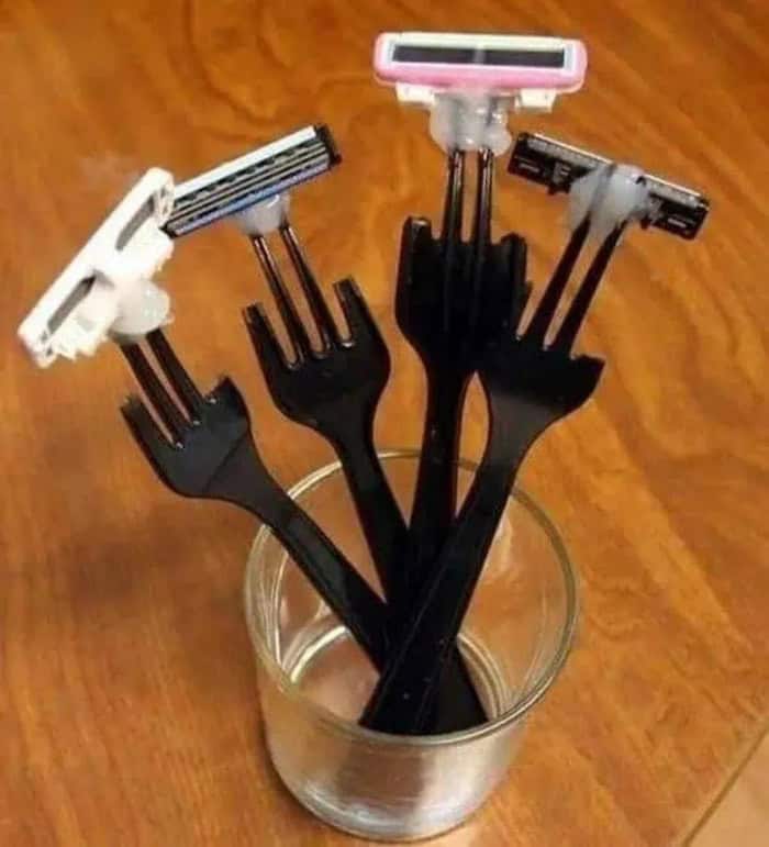 Razor blades attached to black plastic forks in a glass container on a wooden surface.