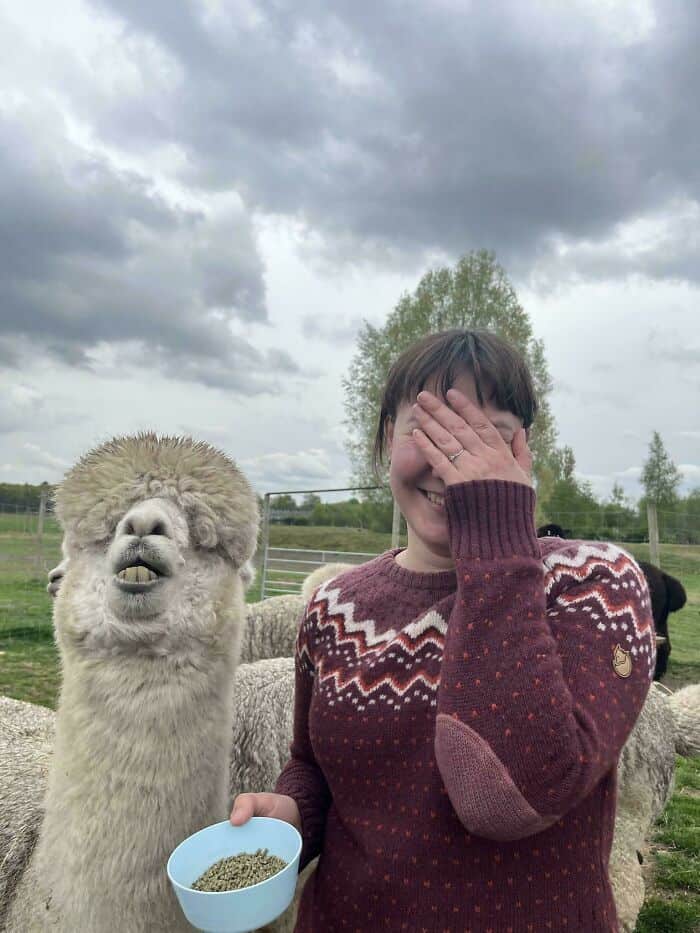 Alpaca and woman enjoying a fun moment outdoors on a farm, with a cloudy sky overhead and lush green.
