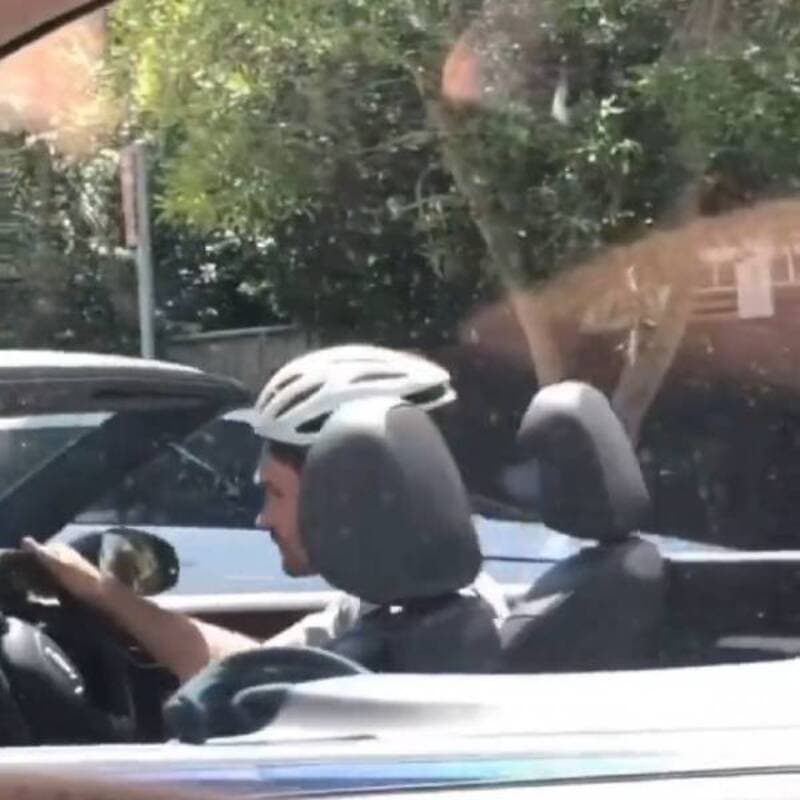 A man wearing a helmet is sitting in a convertible car, looking at his phone, with trees and sunligh.