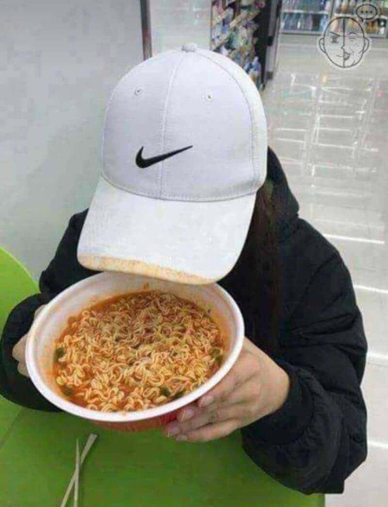 Person with a white Nike cap covering their face, holding a bowl of ramen noodles in a store aisle.