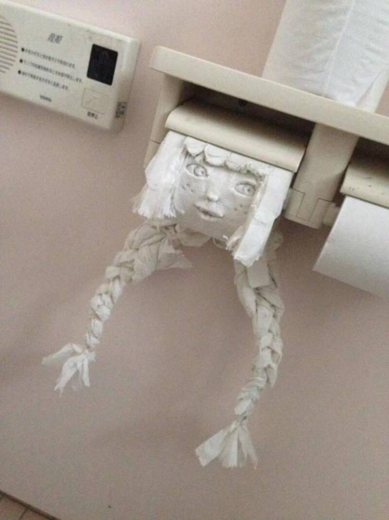 Toilet paper roll with a face drawing and braided paper hair hanging from a bathroom paper holder.