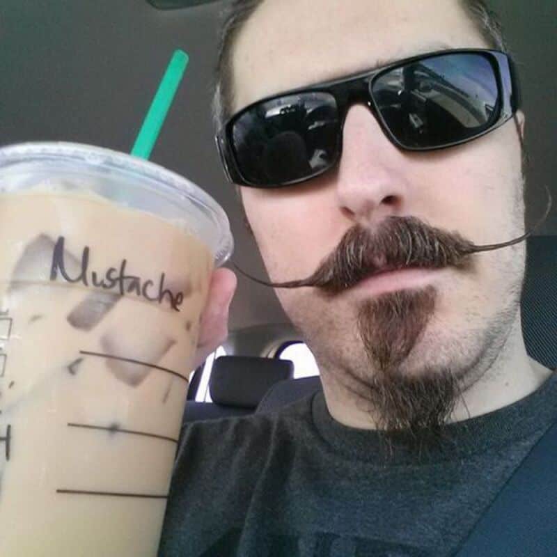 Man with a mustache and beard holding iced coffee in a car, wearing sunglasses.